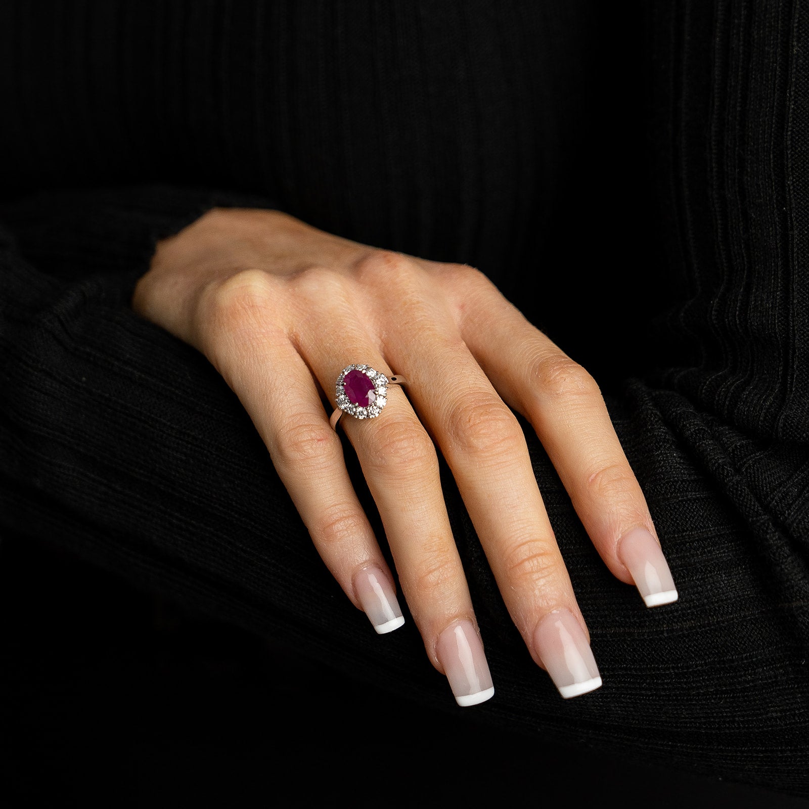 A model wearing a black ribbed sweater showcases a bespoke oval-cut ruby ring. The vibrant red center stone is encircled by a brilliant halo of round-cut diamonds and set on a polished silver-toned band.