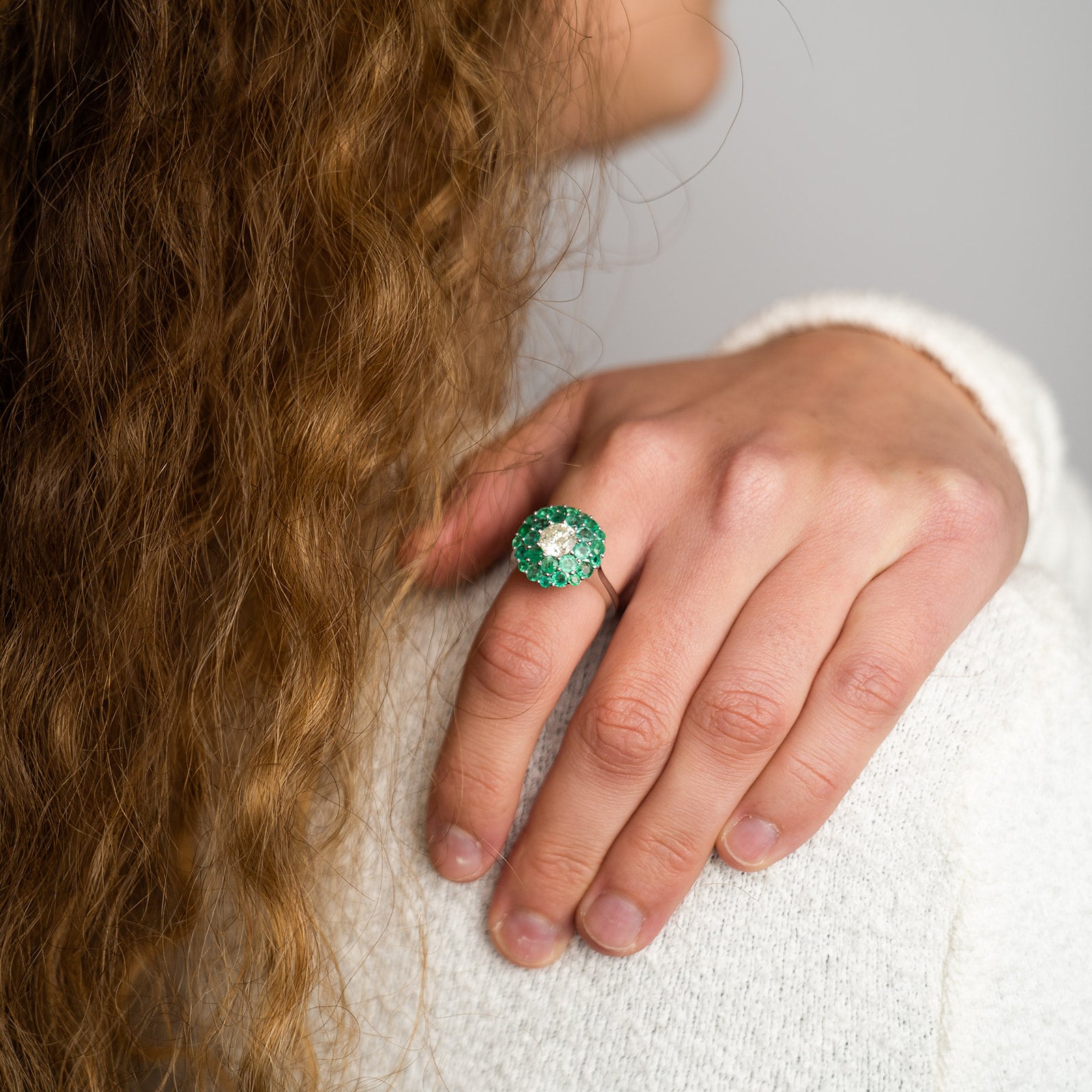 A model displays a vibrant emerald cluster ring featuring a brilliant round diamond center surrounded by two tiers of small, round-cut emeralds.