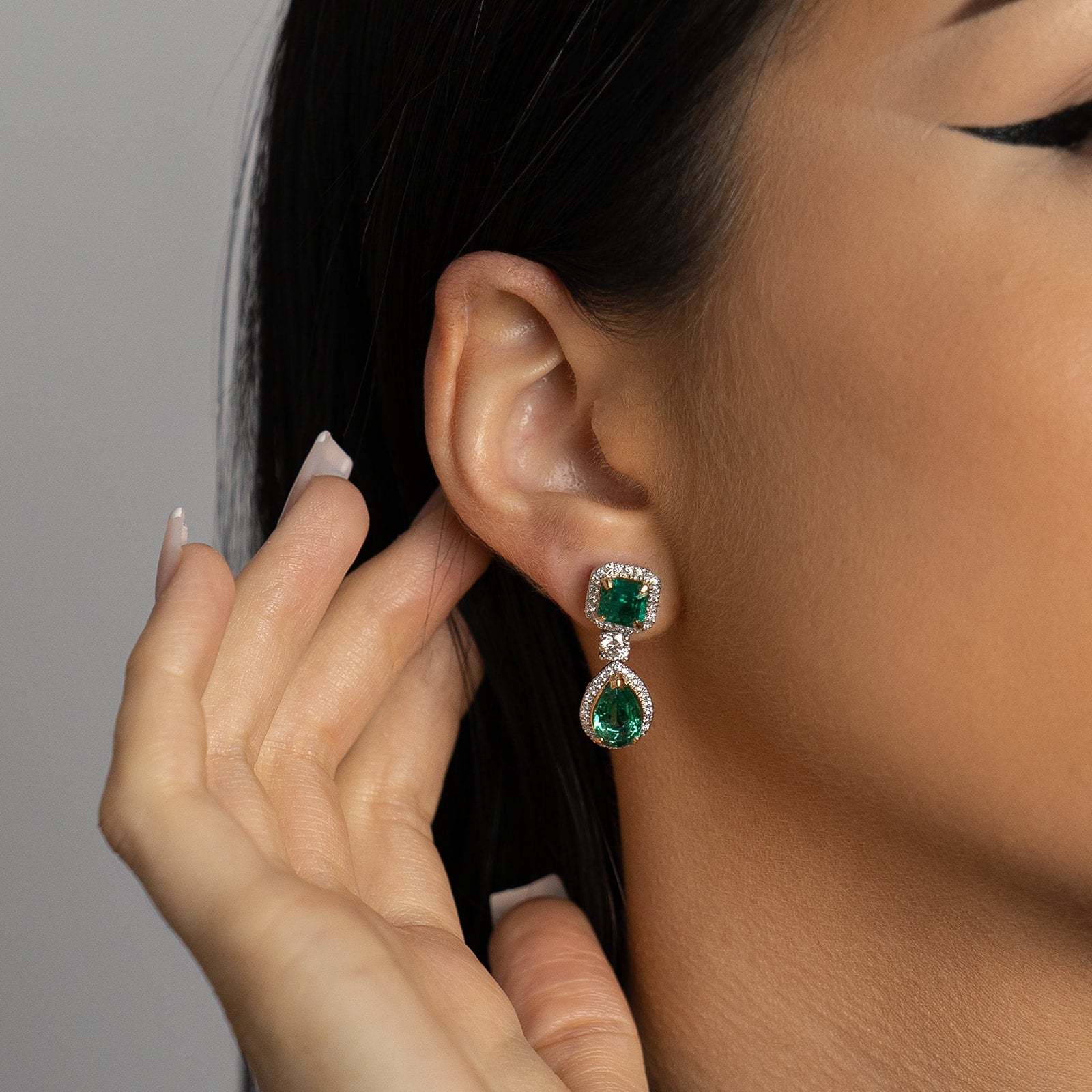 A pair of geometric emerald drop earrings, featuring a square-cut emerald top and a pear-cut emerald drop, both encircled by delicate diamond halos.