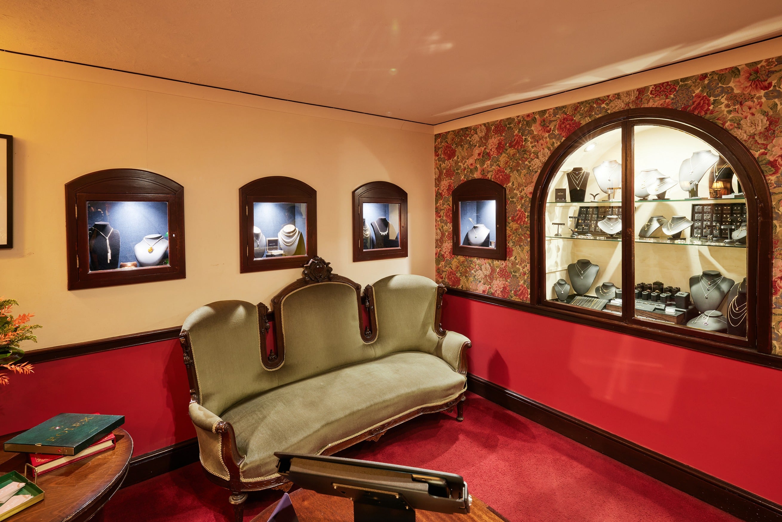 The elegant waiting area of the Ebony Jewellers showroom featuring a vintage green velvet sofa. The walls are decorated with classic floral wallpaper and small, wood-framed jewellery display boxes set into the cream-coloured upper walls.