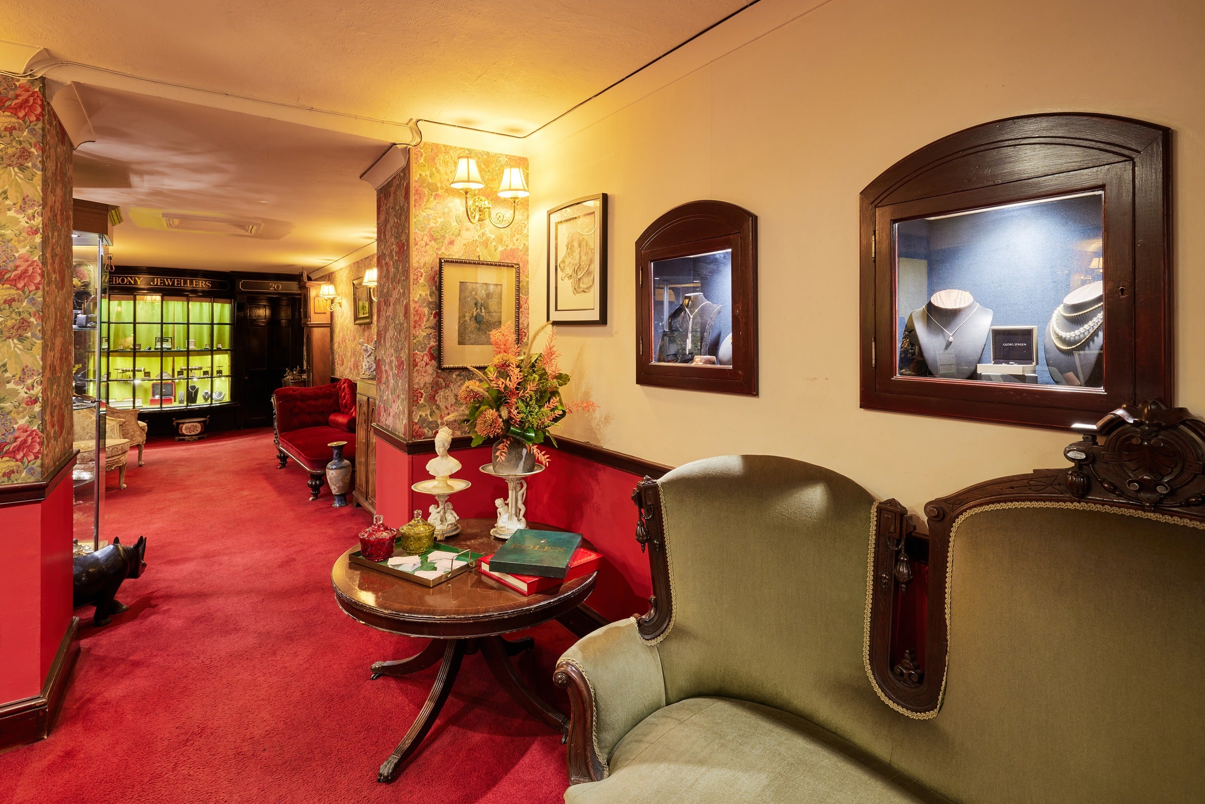 A perspective view of the Ebony Jewellers showroom hallway, highlighting a deep red carpet and walls adorned with floral patterns. Ornate furniture, including an antique side table with decorative glassware and a green velvet armchair, adds to the shop's traditional charm.