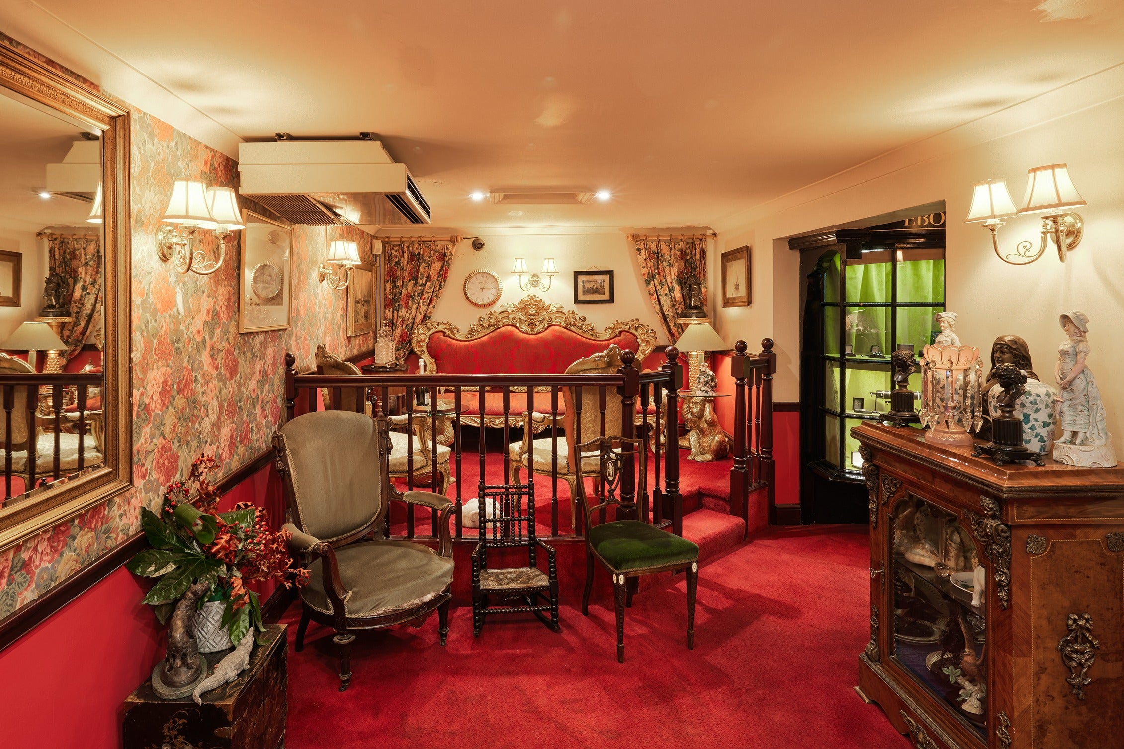 A raised seating lounge within the showroom featuring a magnificent gold-trimmed red velvet sofa. The space is warmly lit by wall-mounted lamps and decorated with antique paintings and rich floral curtains.