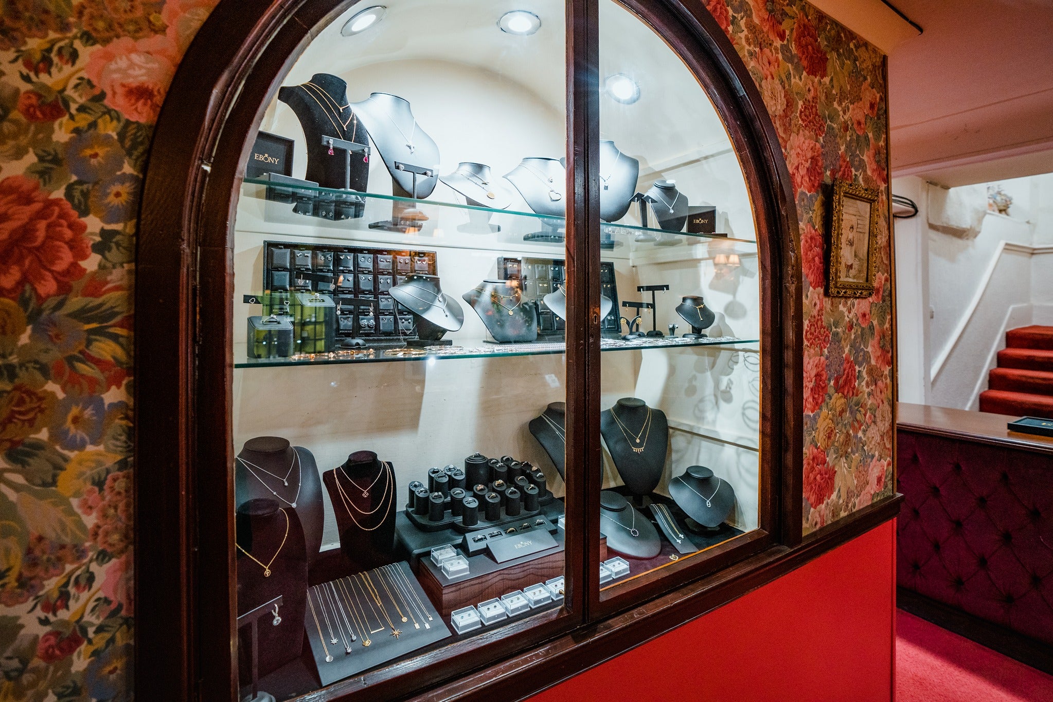 A detailed look at a large, wood-framed arched wall display. Three glass shelves are meticulously arranged with velvet busts showcasing gold necklaces, alongside tiers of rings and earring sets.