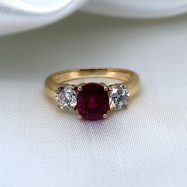 A stunning 18ct yellow gold three-stone ring featuring a vibrant oval-cut ruby surrounded by two sparkling round brilliant diamonds.