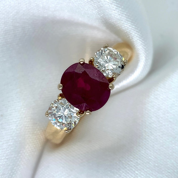 A stunning 18ct yellow gold three-stone ring featuring a vibrant oval-cut ruby surrounded by two sparkling round brilliant diamonds.