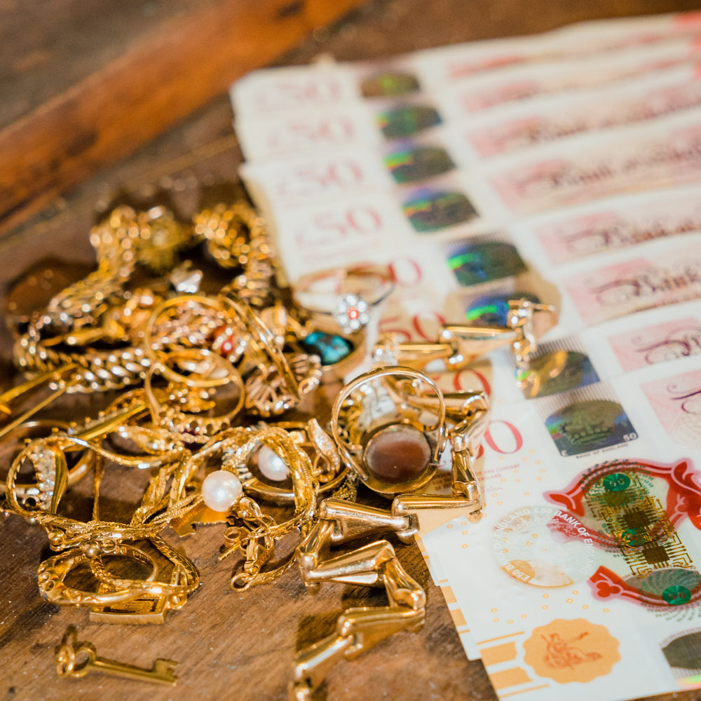 A pile of scrap gold jewellery, including tangled chains, gold rings, and pearl earrings, sits next to a stack of £50 British banknotes. This image represents Ebony Jewellers' service of buying unwanted or broken jewellery for instant cash or bank transfer.