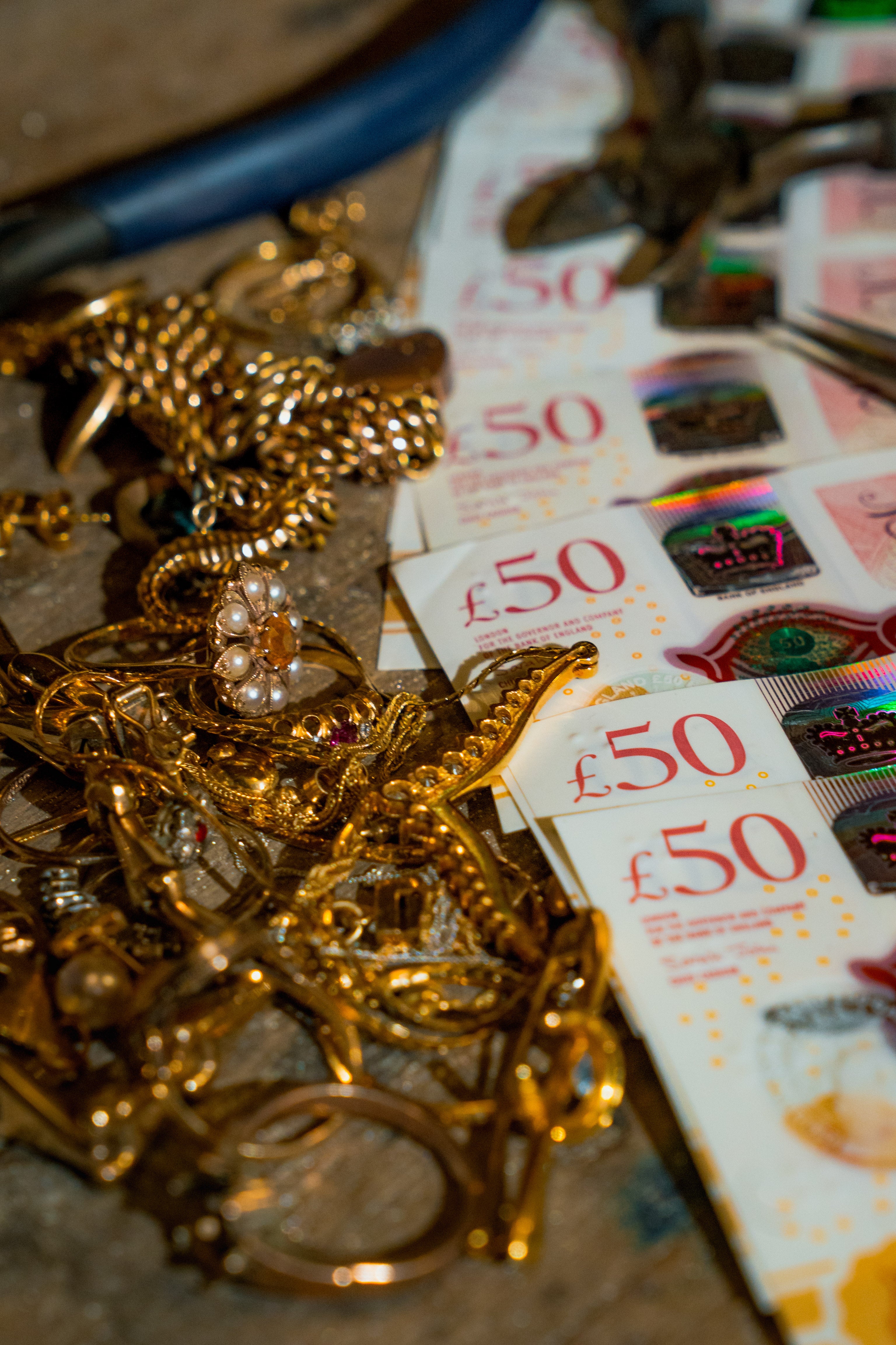 A scattered pile of assorted gold jewellery, including necklaces, rings, and pearl earrings, sits next to a fan of £50 British banknotes. This visual highlights Ebony Jewellers' service of buying unwanted gold for immediate cash or bank transfer.