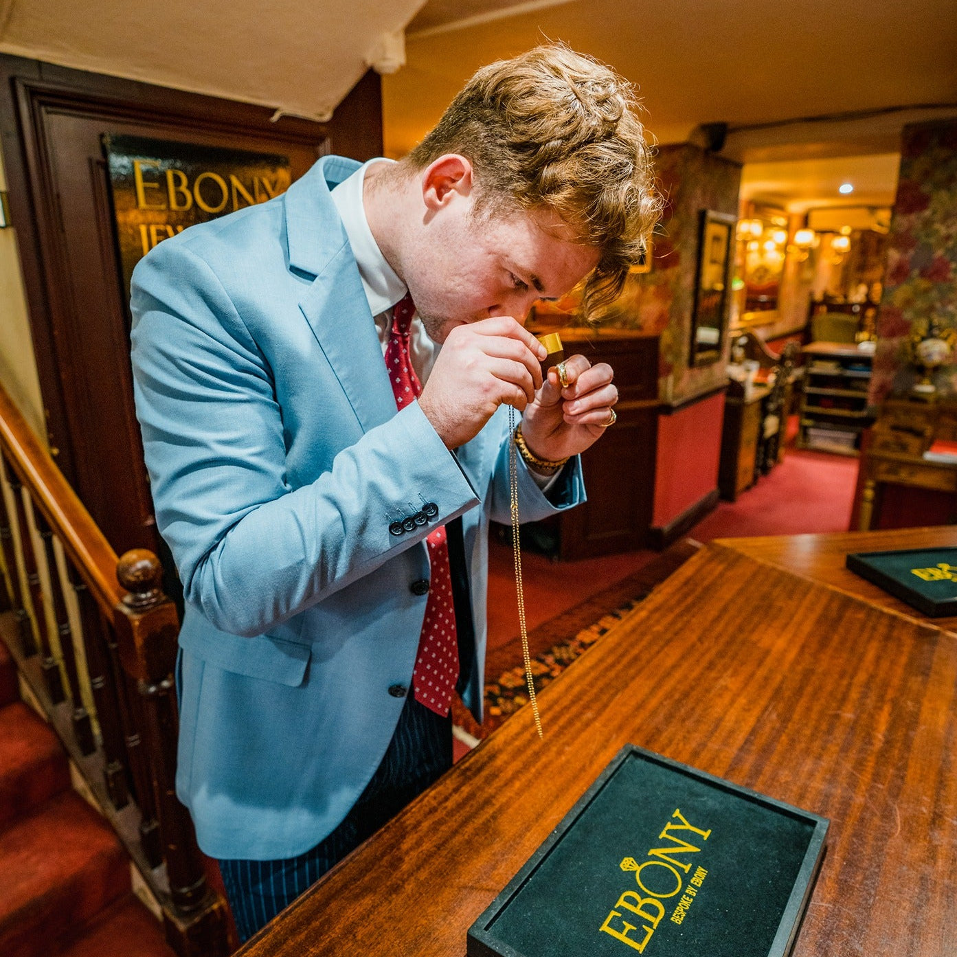 An expert jeweller (Cian Griffin) at Ebony Jewellers in Chichester uses a loupe to inspect a gold ring. He stands behind a wooden counter next to a display tray with the "Ebony" logo, providing a professional and transparent on-the-spot valuation.