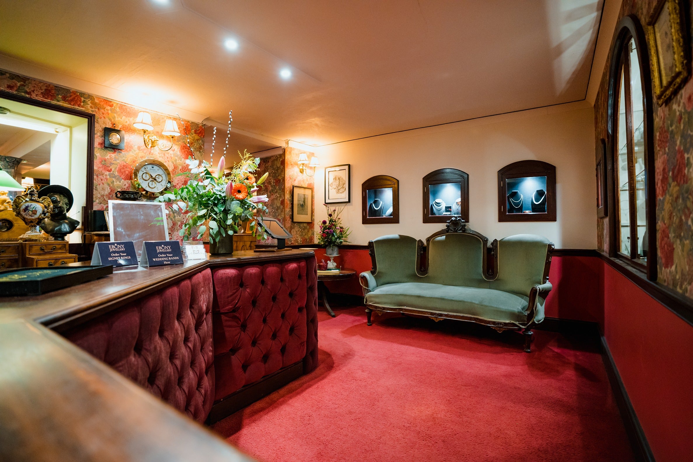 A view from behind the service counter looking toward the seating area. The counter is upholstered in rich red velvet, and a fresh bouquet of flowers sits near the "Signet Rings" and "Wedding Bands" service signs.