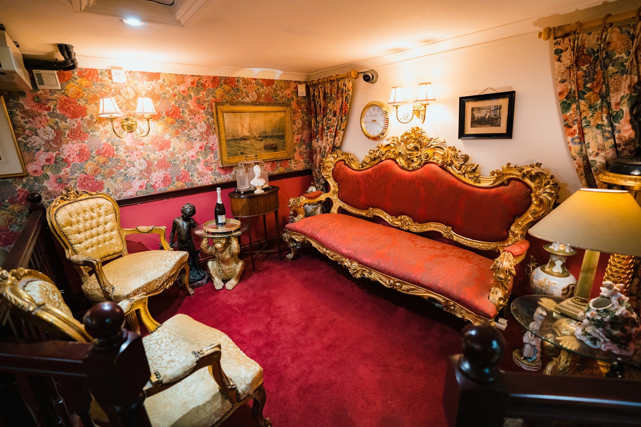 The opulent lounge area featuring a grand, ornate red and gold settee, antique side tables with champagne service, and a classic Rolex-style wall clock.