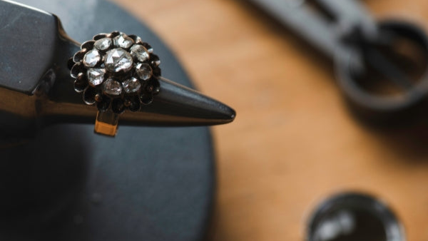 A close-up of a vintage diamond cluster ring secured on a jeweller's mandrel for stone resetting. The piece features a central rose-cut diamond surrounded by a floral halo of smaller diamonds in a darkened silver or antique setting.